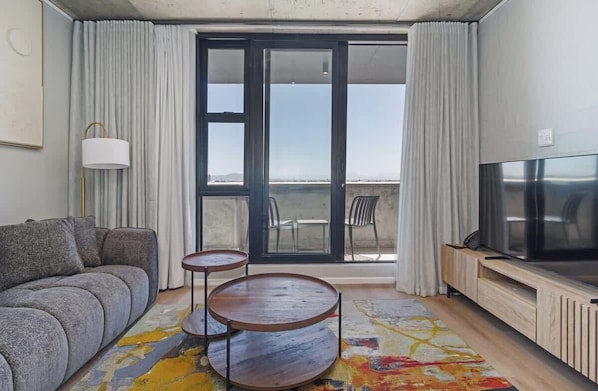 Ein Wohnzimmer mit einer Couch, zwei kleinen Tischen, einem Fernseher und Meerblick durch ein großes Fenster.