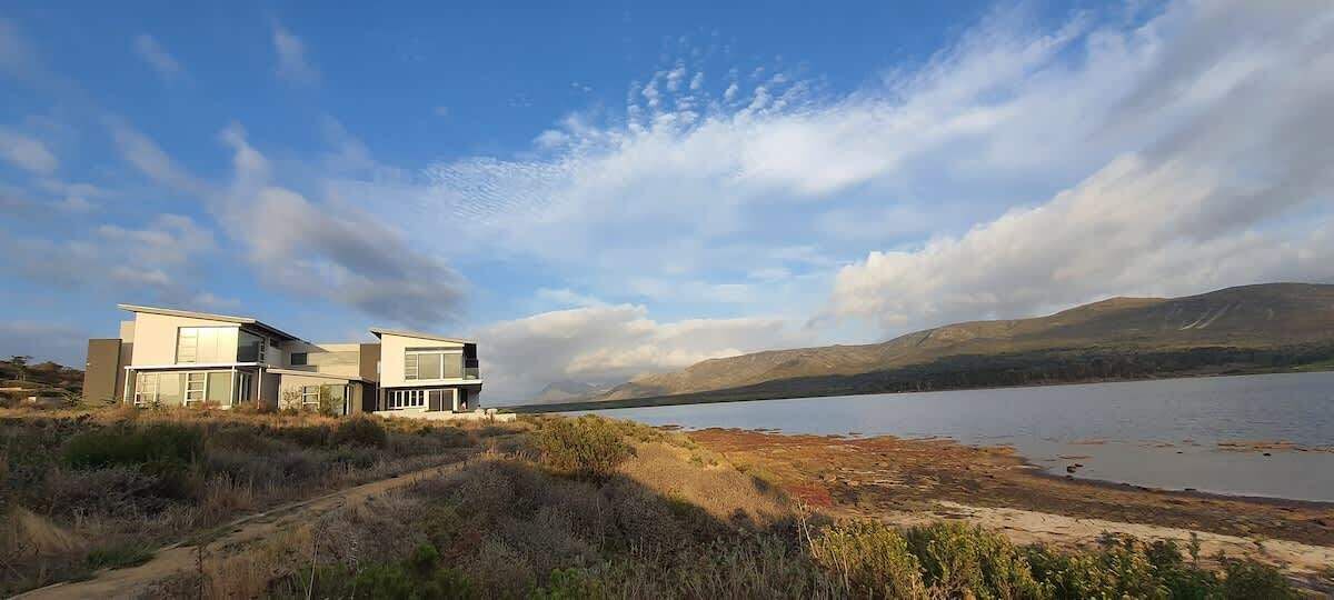 A modern two-story house with large windows, situated near a lake with a clear sky.