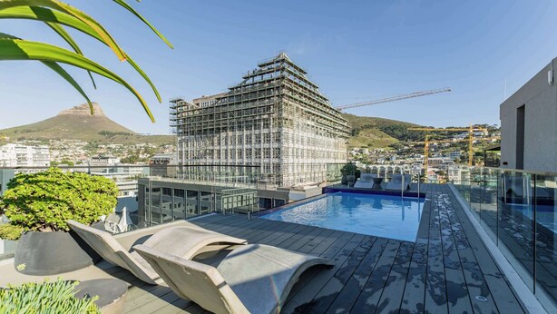 Une piscine sur le toit, agrémentée de chaises longues, offrant une vue sur un chantier de construction et sur une chaîne de montagnes au loin.