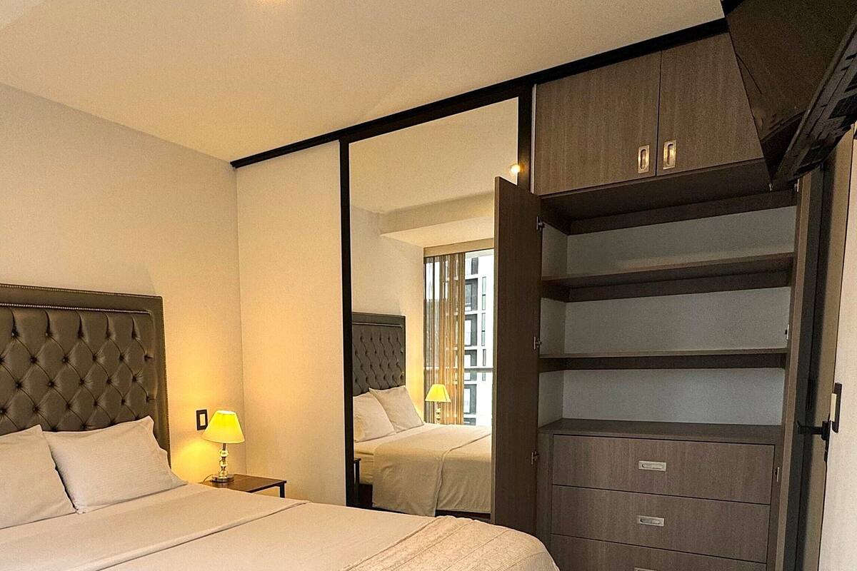 A hotel room with a large bed, a nightstand with a lamp, a wardrobe with multiple drawers, and a mirror reflecting the room.