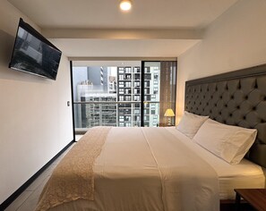 A hotel room with a large bed, a television on the wall, and a view of high-rise buildings through the window. - Aparts4Days 1 Bedroom Apartment 548 (San Isidro)
