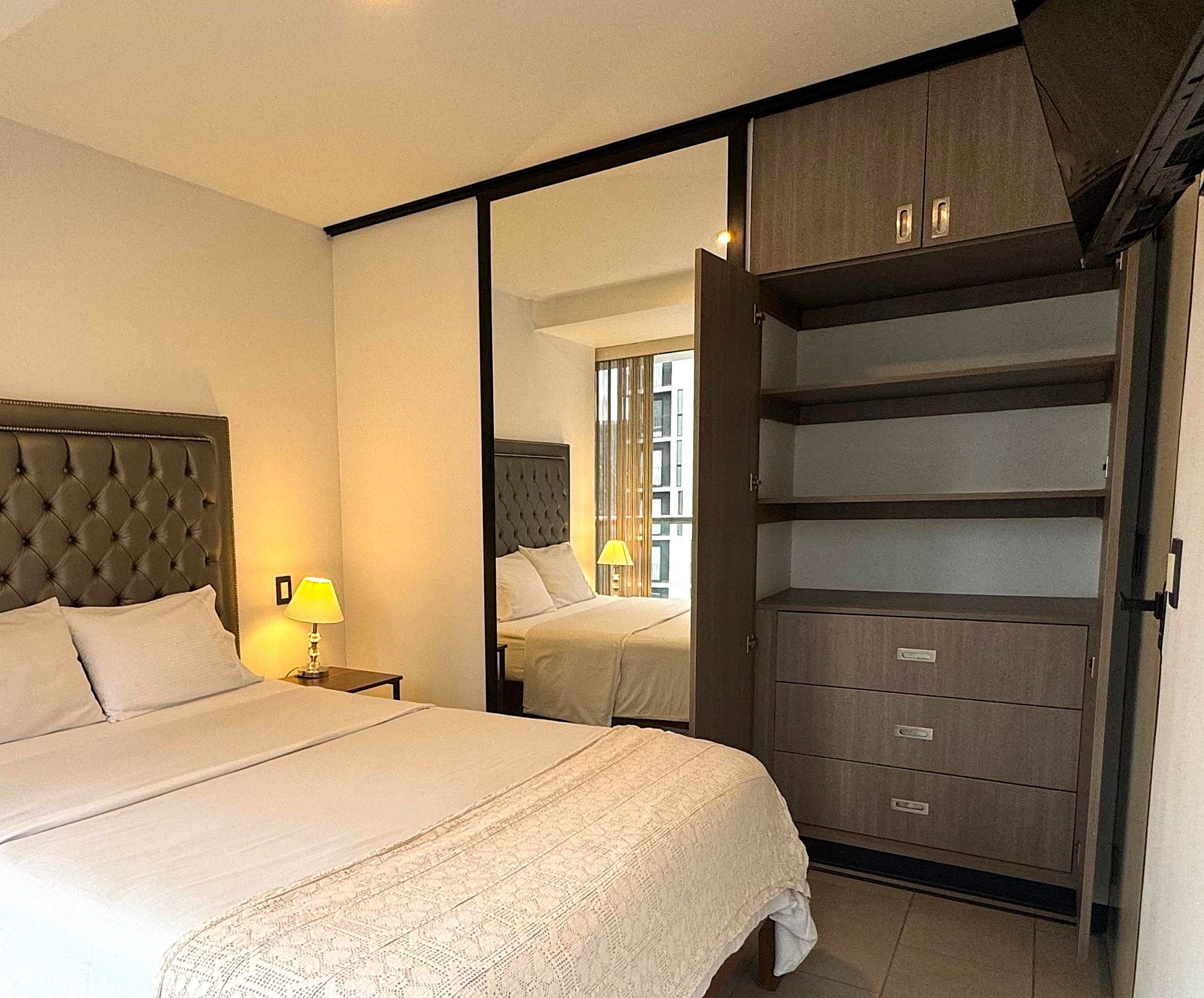 A hotel room with a large bed, a nightstand with a lamp, a wardrobe with multiple drawers, and a mirror reflecting the room.