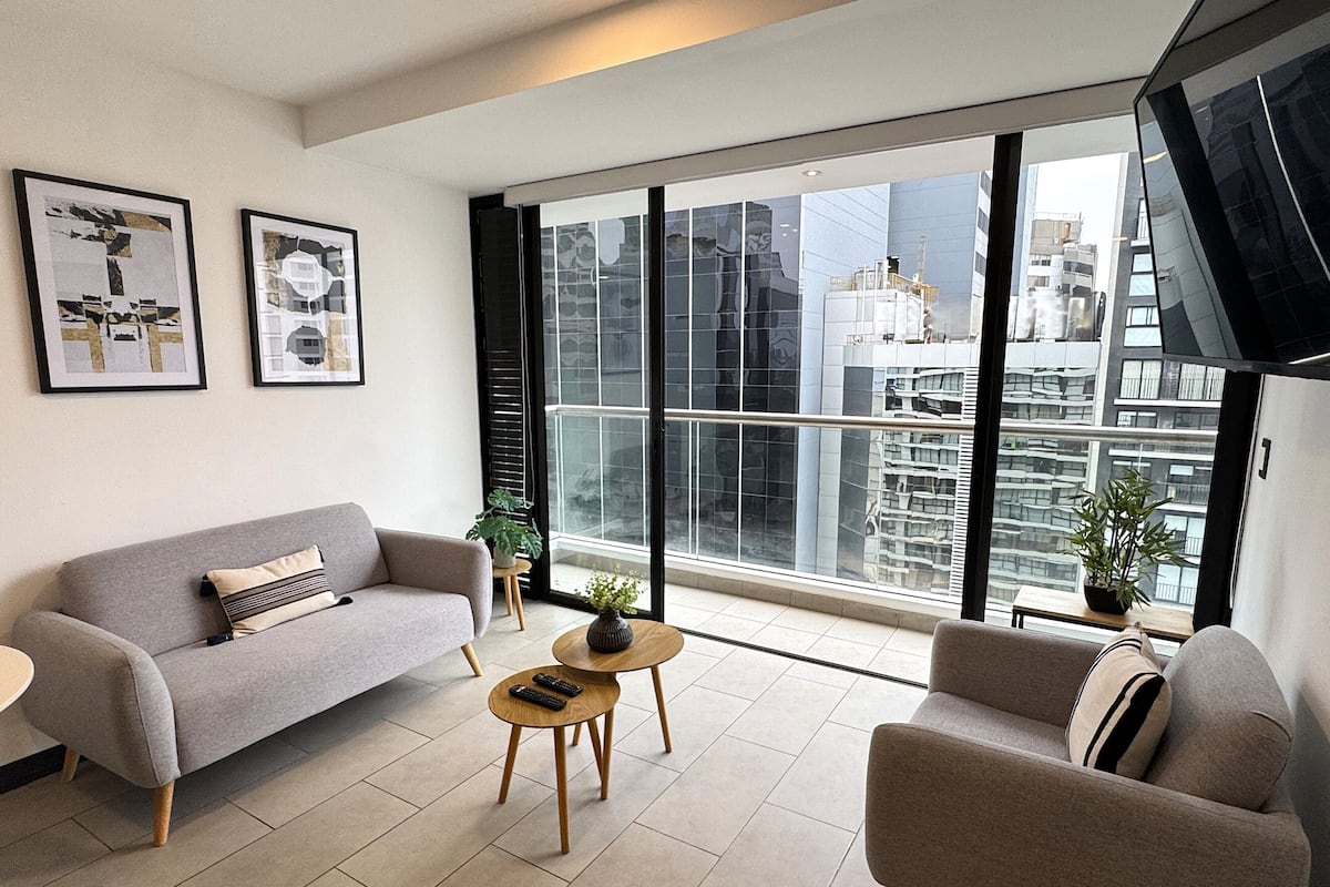 Una sala moderna con un sofá gris, un sillón más pequeño y una mesa de centro. Hay dos cuadros enmarcados en la pared, una planta en maceta sobre una mesita y un ventanal con vistas a la ciudad.