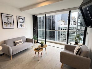 A modern living room with a grey sofa, a smaller armchair, and a coffee table. There are two framed pictures on the wall, a potted plant on a small table, and a large window with a city view. - Aparts4Days 1 Bedroom Apartment 548 (San Isidro)