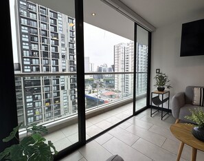 A modern apartment with large windows, a grey sofa, a small wooden table, and a potted plant. - Aparts4Days 1 Bedroom Apartment 548 (San Isidro)