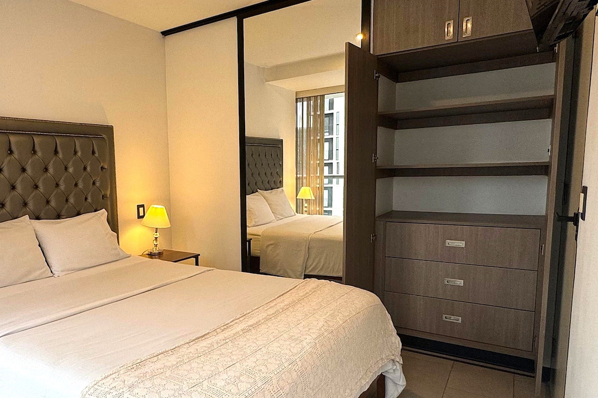 A hotel room with a large bed, a nightstand with a lamp, a wardrobe with multiple drawers, and a mirror reflecting the room.