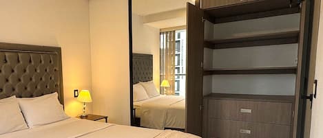 A hotel room with a large bed, a nightstand with a lamp, a wardrobe with multiple drawers, and a mirror reflecting the room.