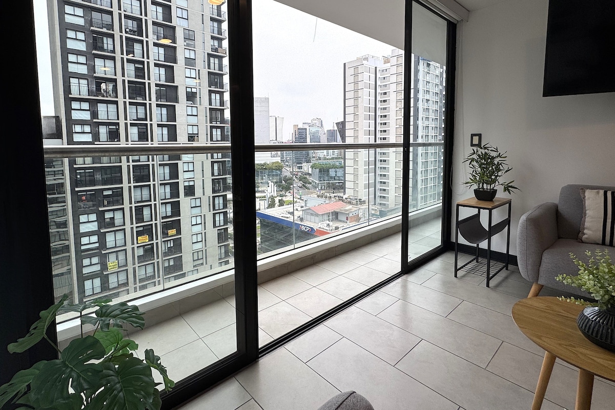 A modern apartment with large windows, a grey sofa, a small wooden table, and a potted plant.
