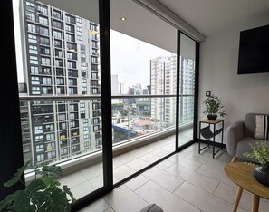A modern apartment with large windows, a grey sofa, a small wooden table, and a potted plant. - Aparts4Days 1 Bedroom Apartment 548 (San Isidro)