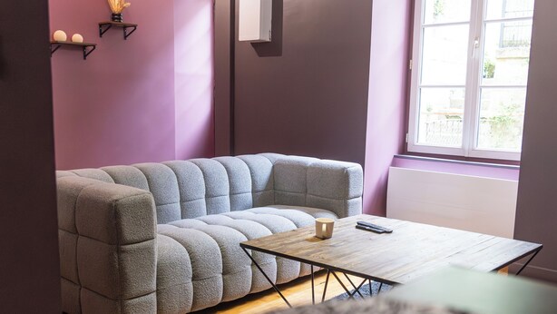 Ein modernes Wohnzimmer mit einem Ecksofa, einem kleinen Holztisch und einem großen Fenster mit Tageslicht.