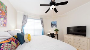 Ein Schlafzimmer mit Bett, Deckenventilator, Fernseher, Schreibtisch mit Stuhl und einem fenster mit Vorhängen.