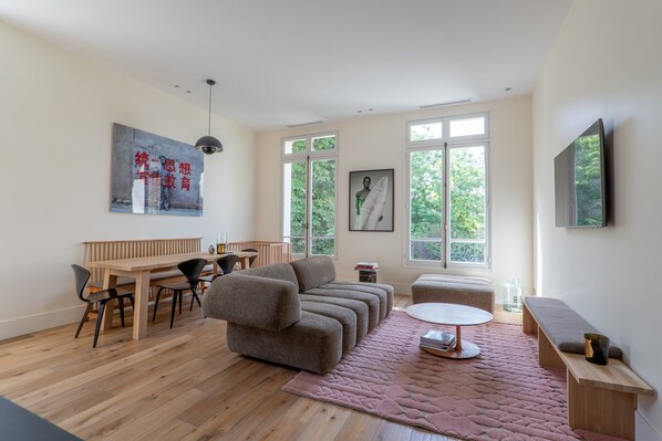 En moderne stue med et træspisebord, en sofa, et rundt sofabord og store vinduer med udsigt til grøn natur.