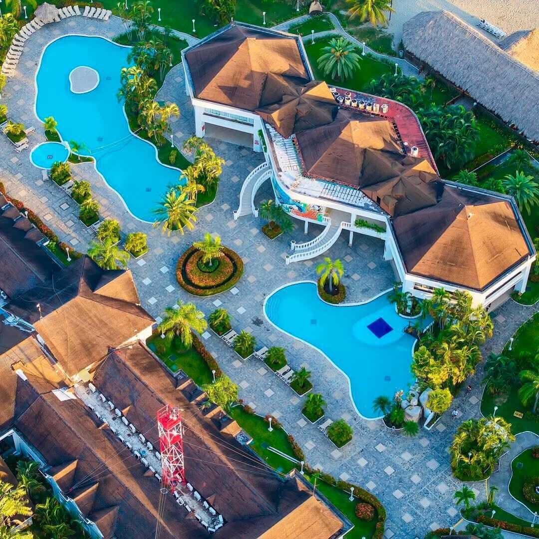 Vue aérienne d’un complexe hôtelier doté de plusieurs piscines, d’un bâtiment central avec un toit incurvé et entouré de palmiers.