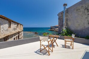 A terrace with two wooden chairs and a table, overlooking a coastal area with a building and a lighthouse. - Skiff - Transformed Studio Sea View (Chania)
