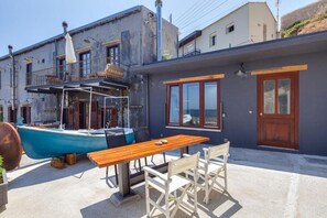 A coastal building with a balcony, a wooden table and chairs, and a blue boat docked outside. - Skiff - Transformed Studio Sea View (Chania)