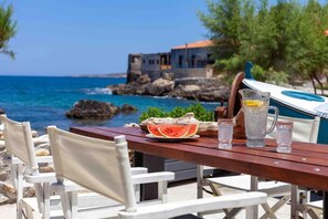 A wooden table with a watermelon and pitchers, overlooking a coastal area with a building and a boat. - Skiff - Transformed Studio Sea View (Chania)