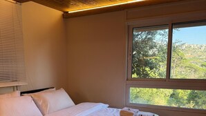 A bedroom with a wooden ceiling, a large bed with white bedding, a window with a view of greenery, and a bedside table with a lamp. - Beel Hills - SOLARA (Mar Mikhael Bnabil)