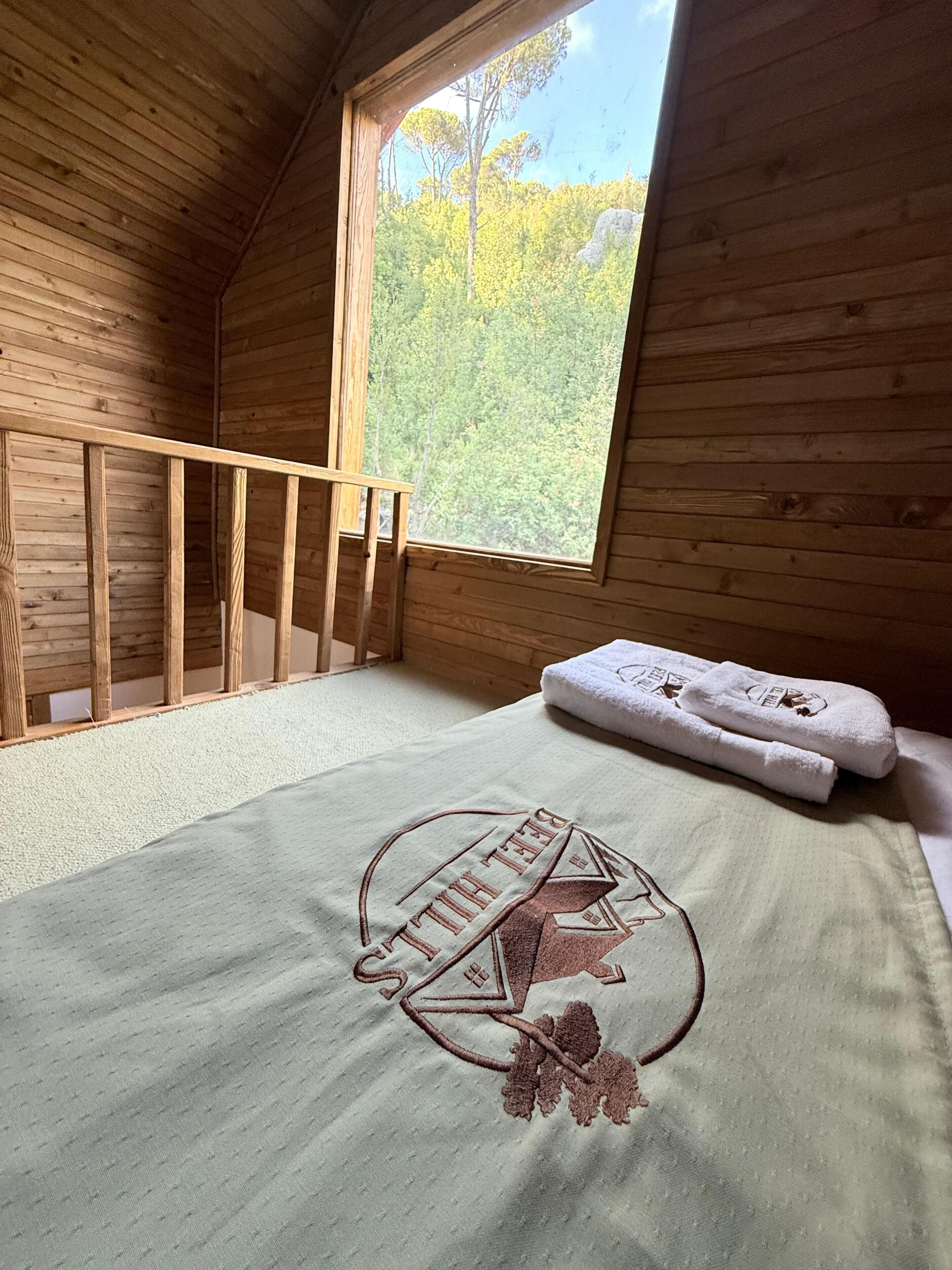 Quarto com paredes de madeira, janela, cama com toalha e prateleira na parede.