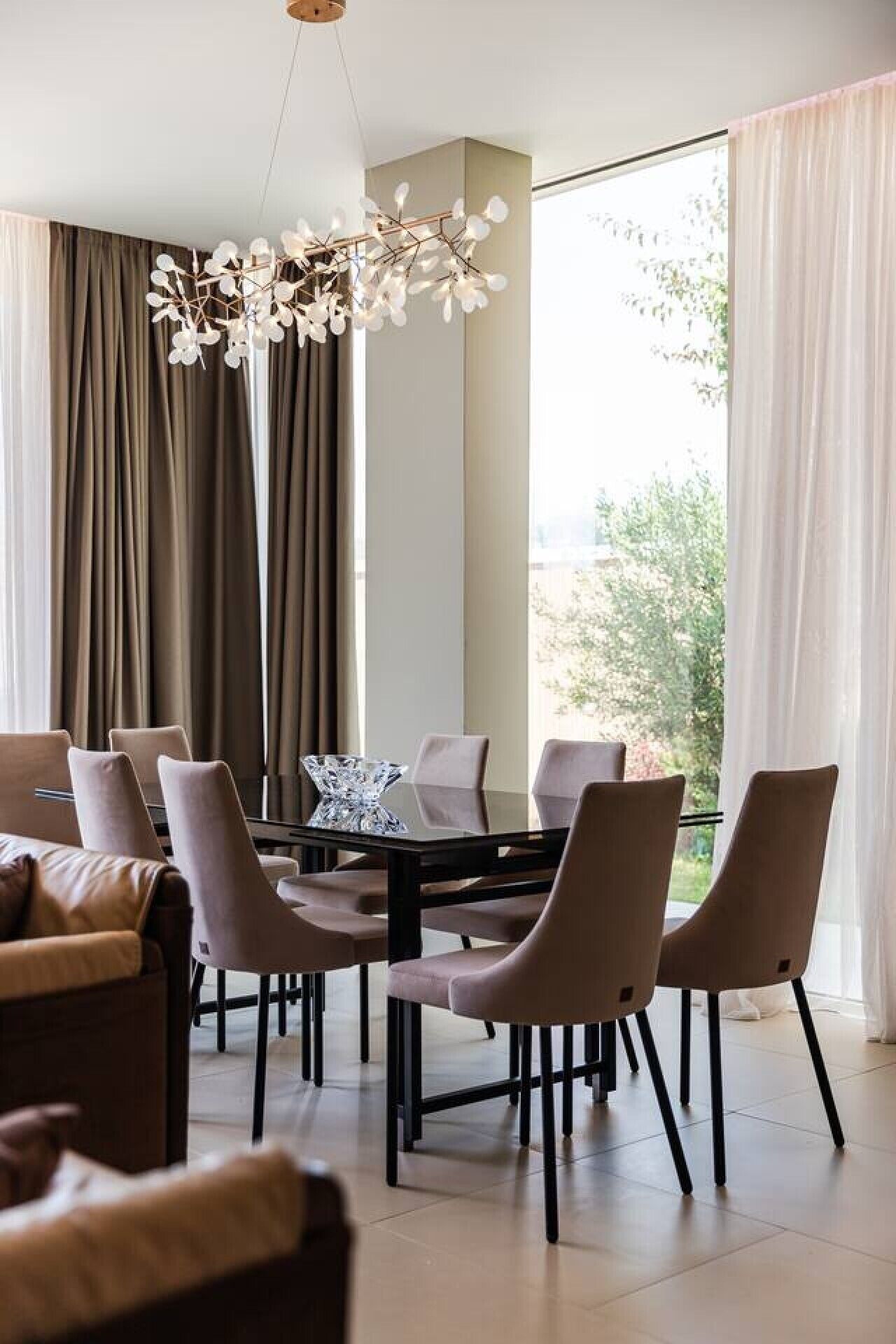 Un comedor moderno con candelabro, ventanas amplias con cortinas y vista a vegetación.