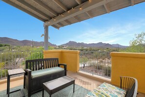 A balcony with a sofa, a small table, and a chair, overlooking a desert landscape with mountains in the distance.
