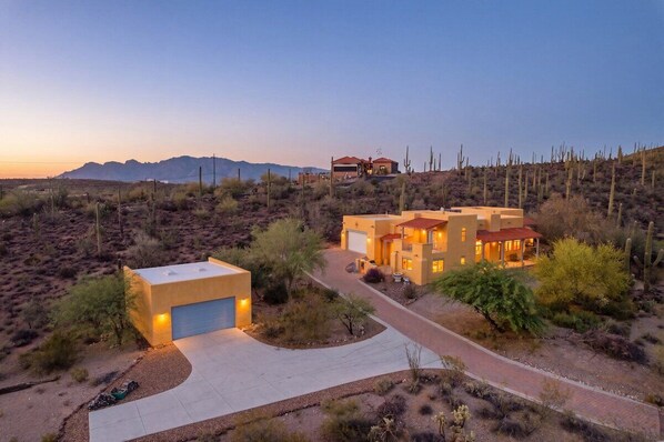 A modern house with a garage, surrounded by desert vegetation and mountains in the background.
