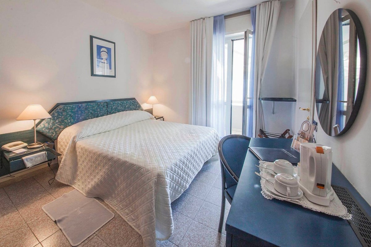 Una habitación de hotel con cama, lámparas de noche, escritorio con cafetera y espejo.