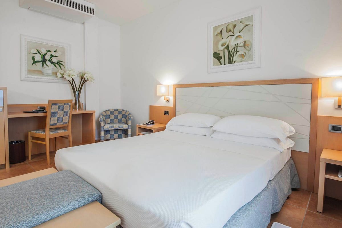 Habitación de hotel con cama, escritorio, silla, mesitas de noche y obras de arte en las paredes.