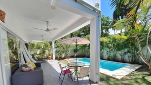 Un espacio exterior techado con piscina, zona de estar con mesa y sillas, y ventilador de techo.