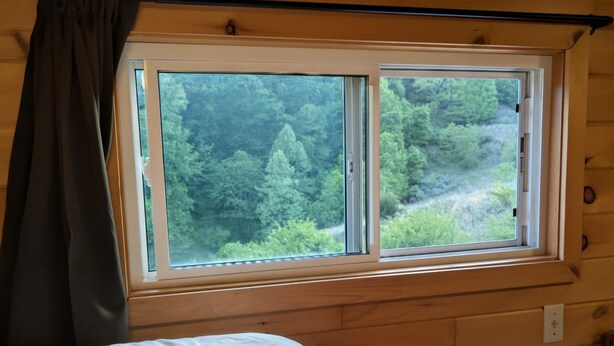 A wooden cabin room with a bed, a window overlooking a forest, and a curtain. - Gorgeous tiny home with mountain views in peaceful Candler (Candler)