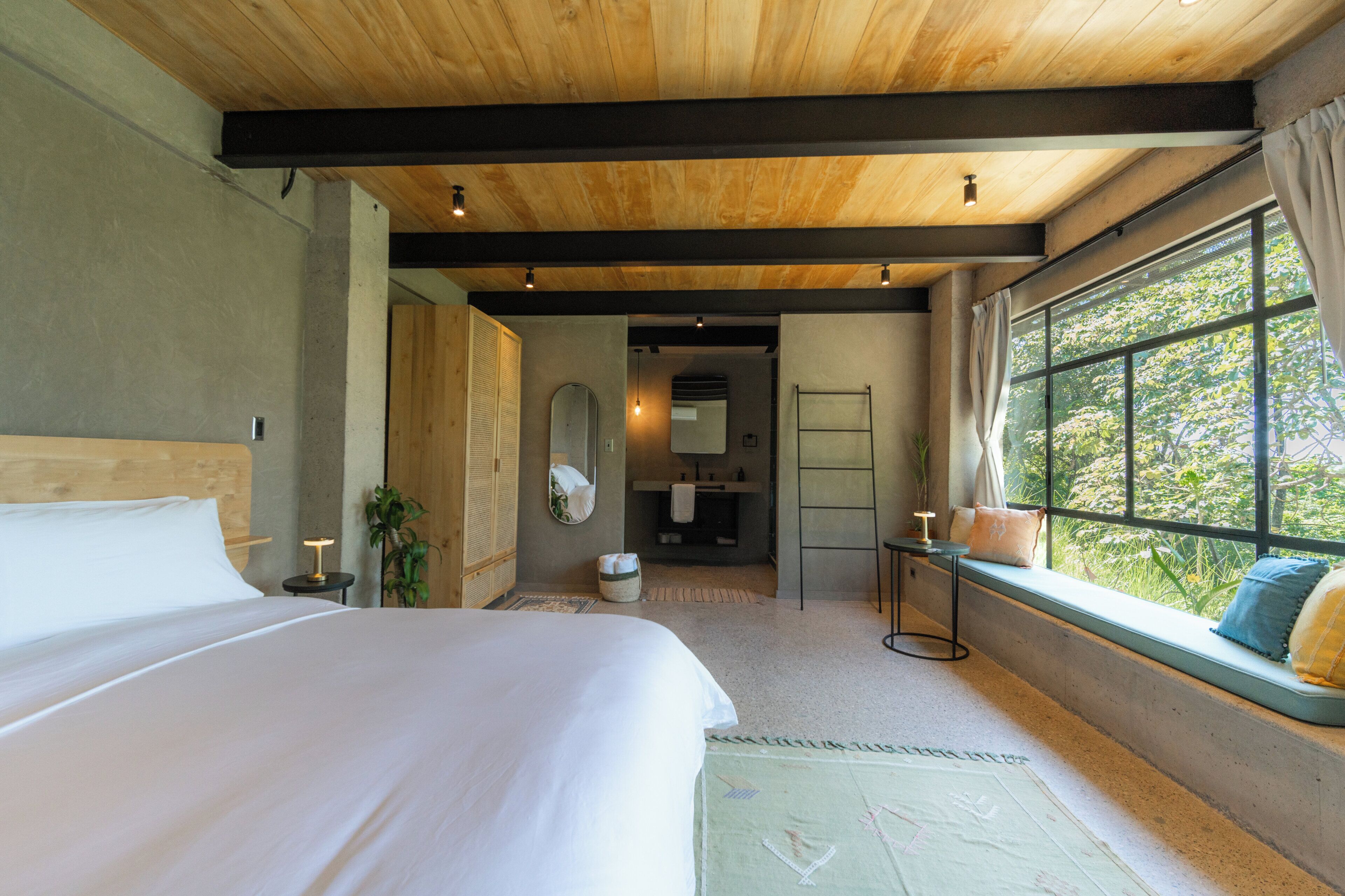 A bedroom with a large bed, a window seat with cushions, a mirror, and a bathroom visible through an open door.