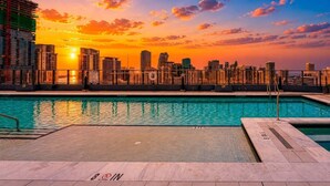 Uma piscina no terraço com o horizonte da cidade ao pôr do sol, um aviso de 'NO DRINKS' e um aviso de 'NO ENTRY'.