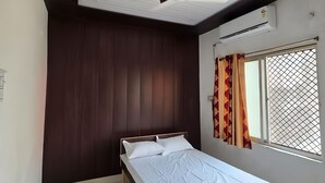 Een slaapkamer met een bed, een plafondventilator, een airconditioningapparaat, een raam met gordijnen en een luchtverkoeler aan de muur.