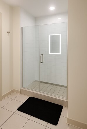 Eine begehbare, gläserne Dusche mit Handlauf, einer schwarzen Badmatte und einer deckeneingebauten Lichtleuchte.
