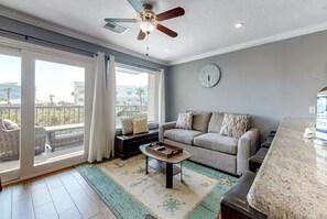 A living room with a sofa, a coffee table, a ceiling fan, a clock, and a view through a sliding door to an outdoor balcony. - 2 Pools, Hot Tub, Dogs Free, Right on the Beach! (Galveston)