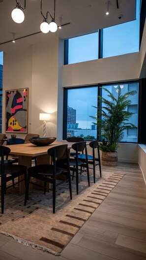 Ein moderner Essbereich mit großem Tisch, Stühlen, einer Stehlampe, einer Zimmerpflanze und Blick auf die Stadt durch große Fenster.