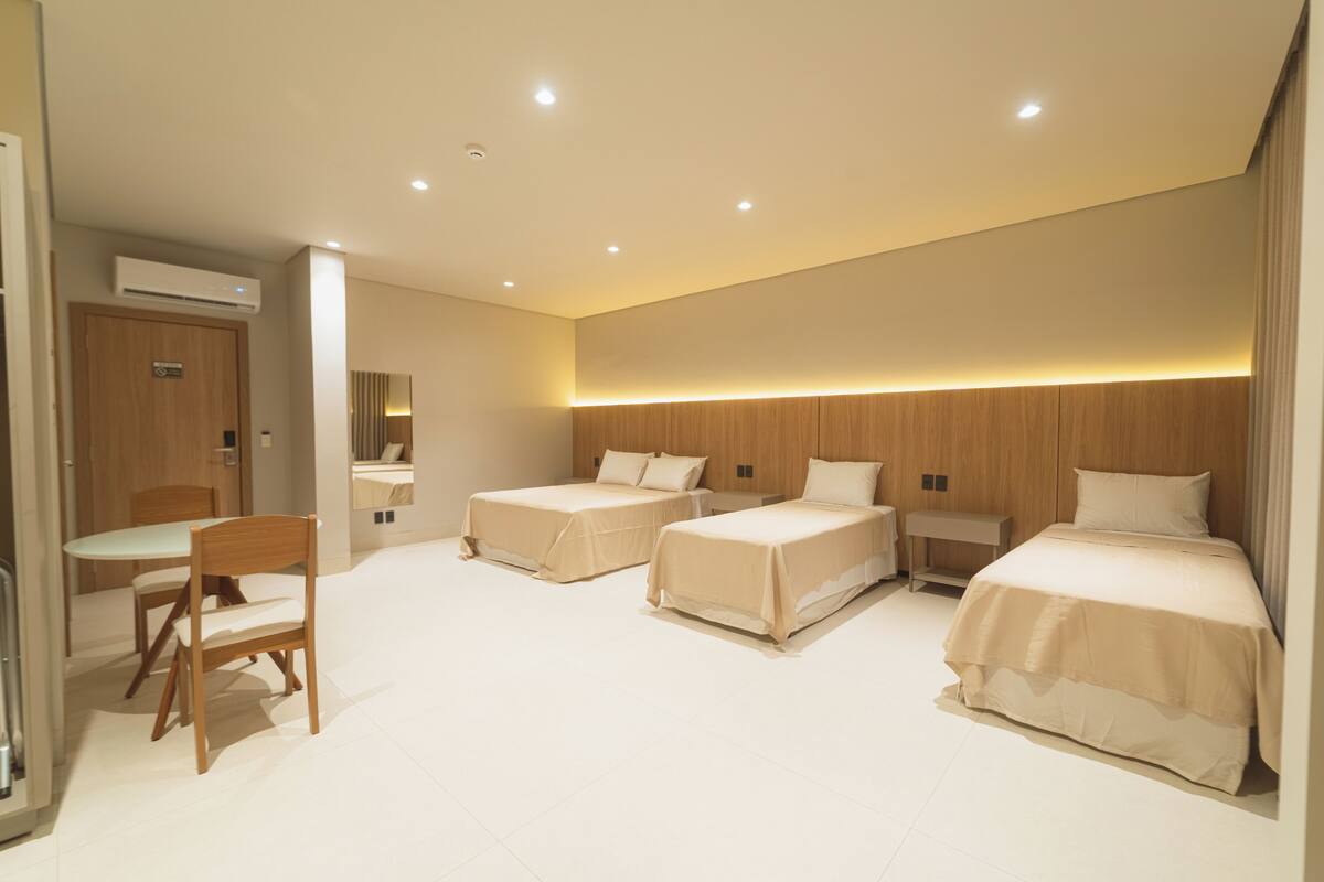 Habitación de hotel con dos camas individuales, una cama doble, una silla de madera, una mesita y un espejo.