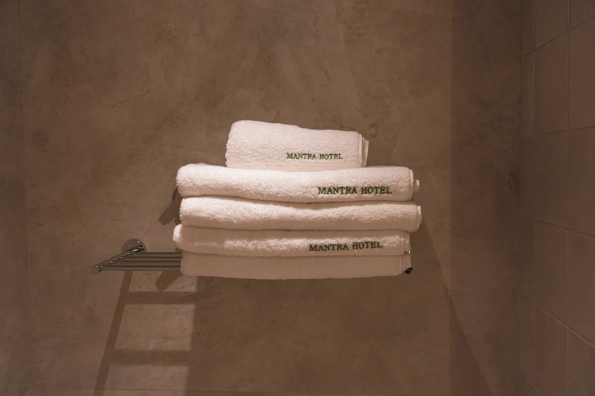 Una pila de toallas blancas con la inscripción 'MANTRA HOTEL', colocadas sobre un perchero metálico en un baño con baldosas color crema.