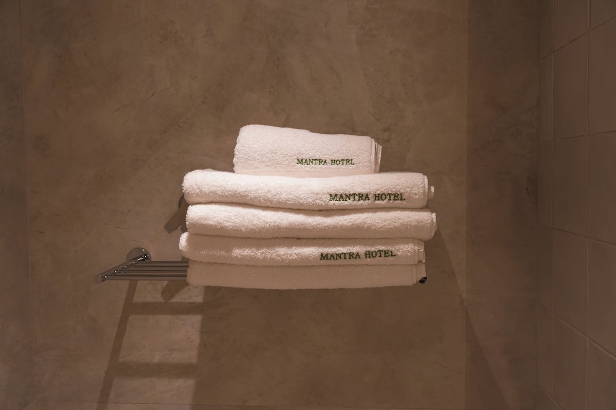 Une pile de serviettes blanches sur lesquelles est imprimé le nom « MANTRA HOTEL », posée sur un porte-serviettes en métal dans une salle de bain carrelée de carreaux beige.