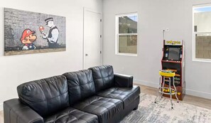 Un salon avec un canapé en cuir noir, un tableau représentant Mario et Bowser, une borne d’arcade Pac-Man et une fenêtre donnant sur le ciel.