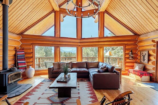 A log cabin interior with a large wooden ceiling, a fireplace, a sofa, and a dining table. - Log Home w/view, Ping Pong, Cornhole, Gas FirePit! (Bend)
