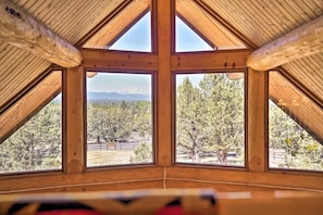 A room with a sloped wooden ceiling and large windows offering a view of a forested area. - Log Home w/view, Ping Pong, Cornhole, Gas FirePit! (Bend)