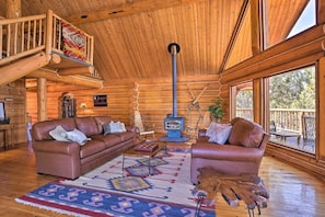 A log cabin interior with a wooden ceiling, two leather sofas, a central wood-burning stove, and a large window overlooking the outdoors. - Log Home w/view, Ping Pong, Cornhole, Gas FirePit! (Bend)