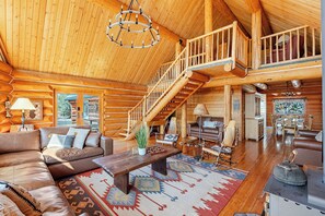 Ein großzügiges Blockhaus-Interieur mit einer zentralen Treppe, einem gemütlichen Wohnbereich mit Sofa und Couchtisch sowie einem Essbereich mit Holzmöbeln.