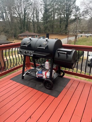 Un barbecue sur une terrasse rouge, avec un sac de charbon de bois et une bouteille d’allume-feu.