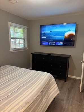 Une chambre à coucher avec un lit, un téléviseur fixé au mur, une commode et une fenêtre avec des stores.