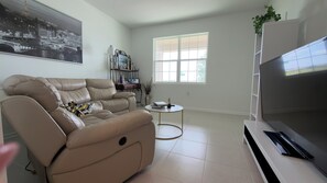 Ein Wohnzimmer mit einem beigen Sofa, einem kleinen runden Tisch, einem Fernseher und einem Fenster mit Jalousien.