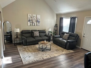 A living room with a dark leather sofa, a coffee table, and a rug. There are two lamps, a mirror, and a painting on the wall.