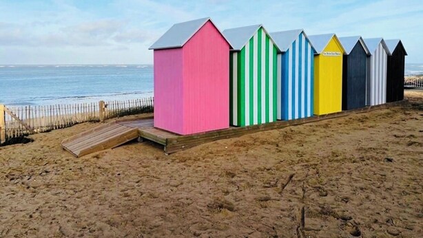 Een rij strandhutten met een houten terras ernaartoe, gelegen op een zandstrand met de zee op de achtergrond.