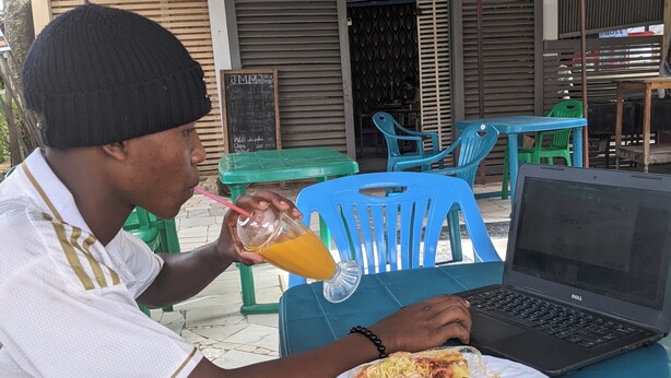A person sitting at a table with a laptop and a plate of food, under a covered outdoor area with chairs and a building in the background. - Anna Luxury Lodge - Arusha  (Arusha)