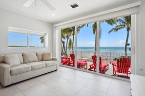 Ein Zimmer mit weißem Sofa, einem Balkon mit roten Stühlen und Blick auf Strand und Meer.